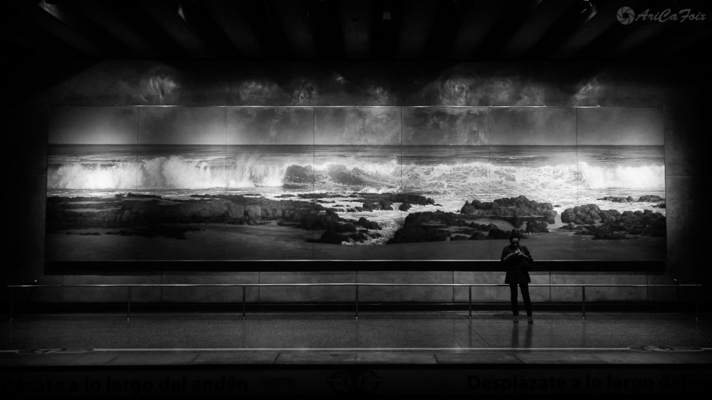 Un hombre espera el Metro, solo en el andén, de pie frente a una pintura del mar.